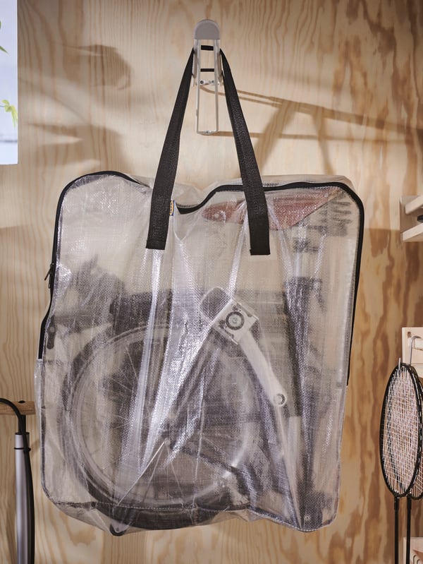 A transparent DIMPA storage bag with a fold-up bicycle in it hanging on a white SKRÄLL hook attached to a wooden wall.