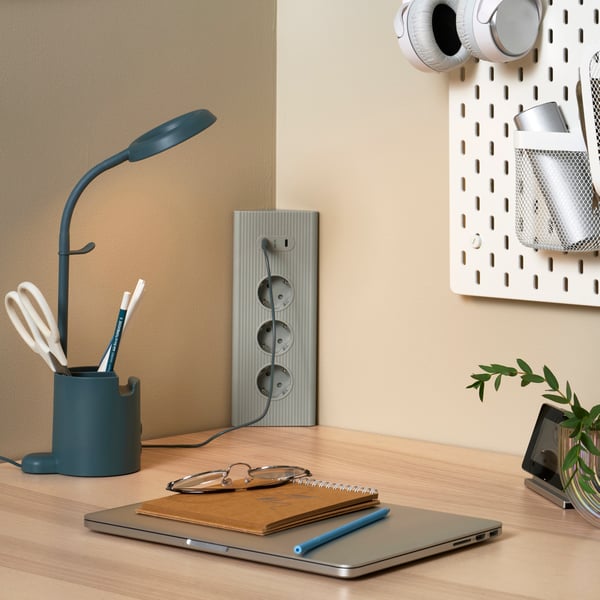 A green SKOTAT extension cord in the corner of a desk and fitted into the corner of a room with a USB cable connected to it.
