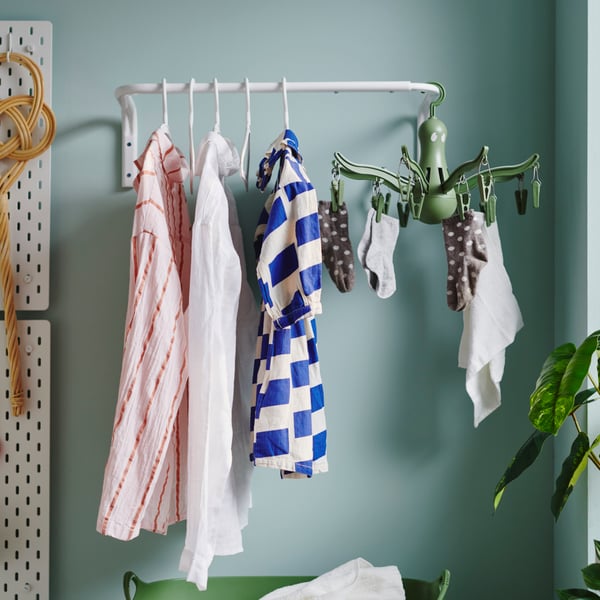 A white, powder-coated MULIG clothes bar is on a light-green wall. It is holding blouses and an octopus-looking hang dryer.