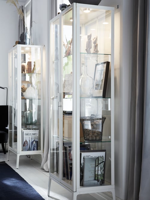 Deux vitrines MILSBO blanches sont placées contre un mur. Elles contiennent des livres, des vases et d’autres objets éclairés.