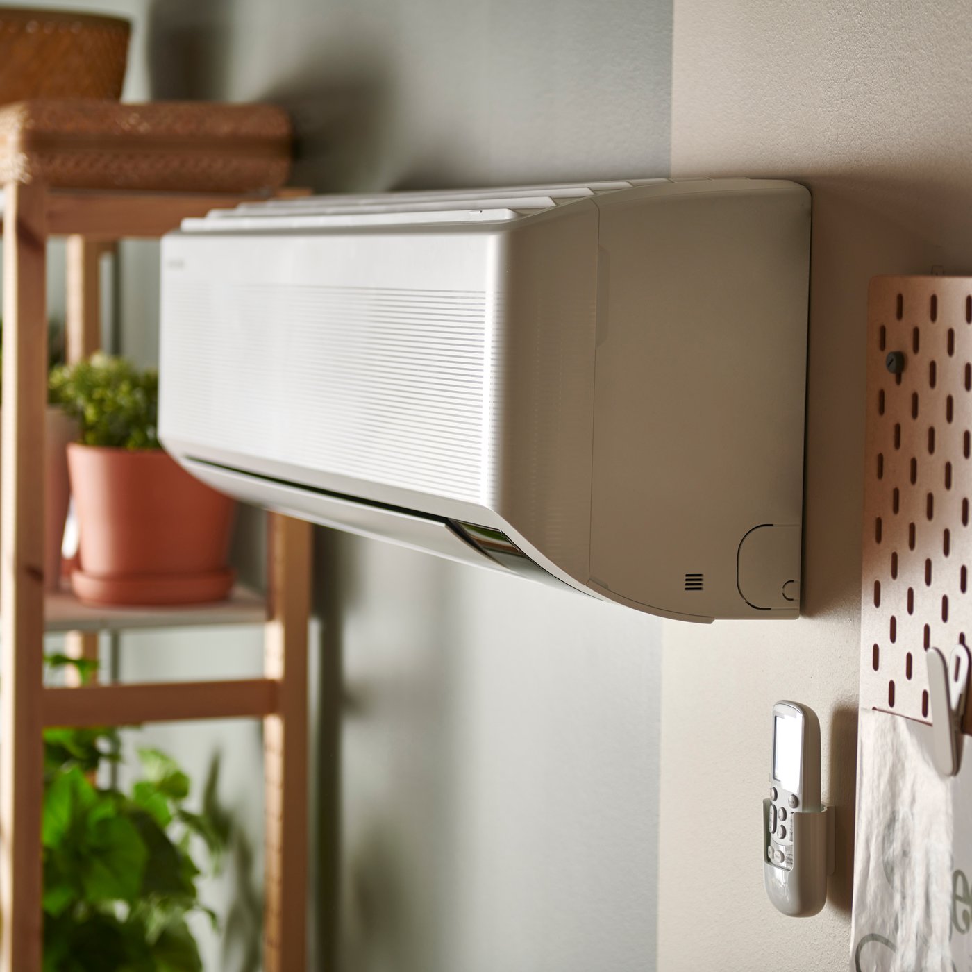 Väggmonterad värmepump i ett rum. Till vänster står en hylla med krukväxter, och till höger syns en pegboard med upphängda föremål.