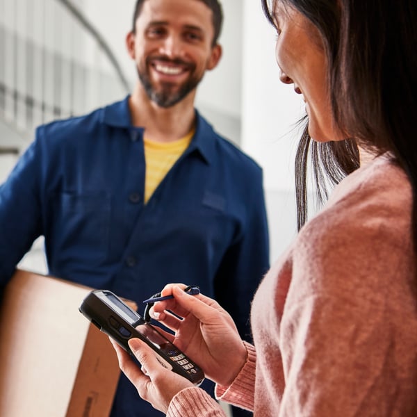 A delivery man in a blue jacket hands something to sign to a woman wearing a grey jumper. Next to her are flatpack boxes and an IKEA blue bag.