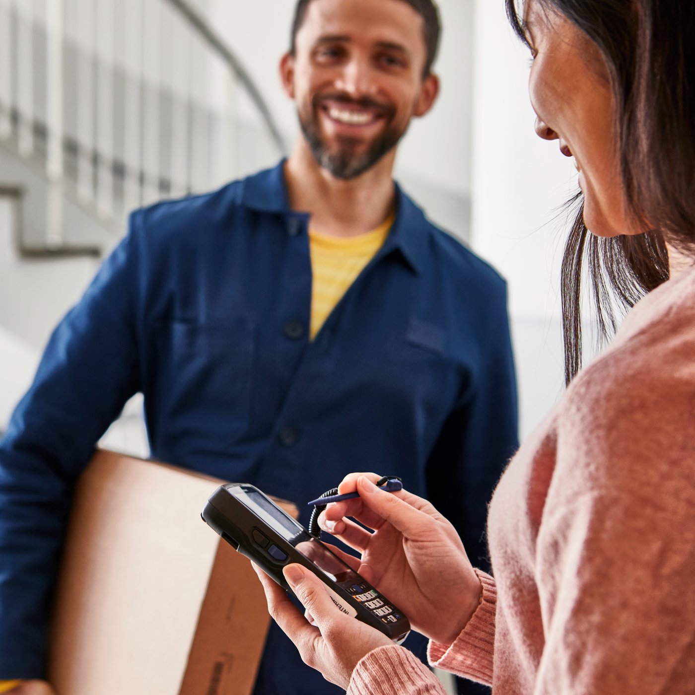 A delivery man in a blue jacket hands something to sign to a woman wearing a grey jumper. Next to her are flatpack boxes and an IKEA blue bag.