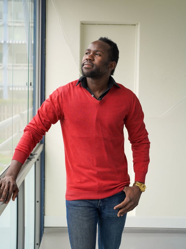 Hassan stands on his balcony in a red sweater, looking out the window.