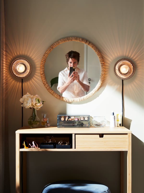 Des appliques en verre SOLKLINT laiton/gris se trouvent de chaque côté d'un miroir naturel sur une coiffeuse en bambou.