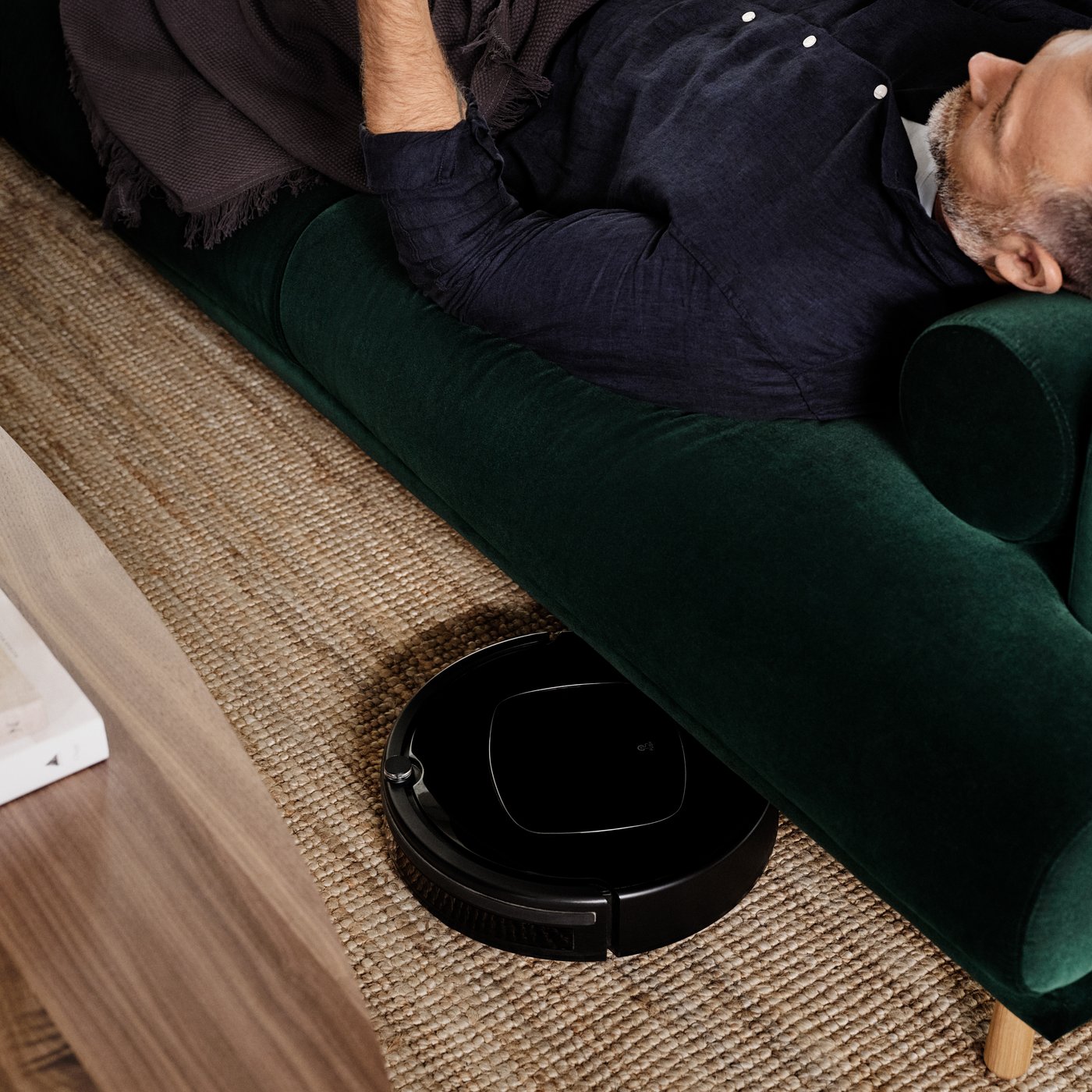 A natural LOHALS rug, flatwoven under a green sofa with a robot vaccum cleaning 