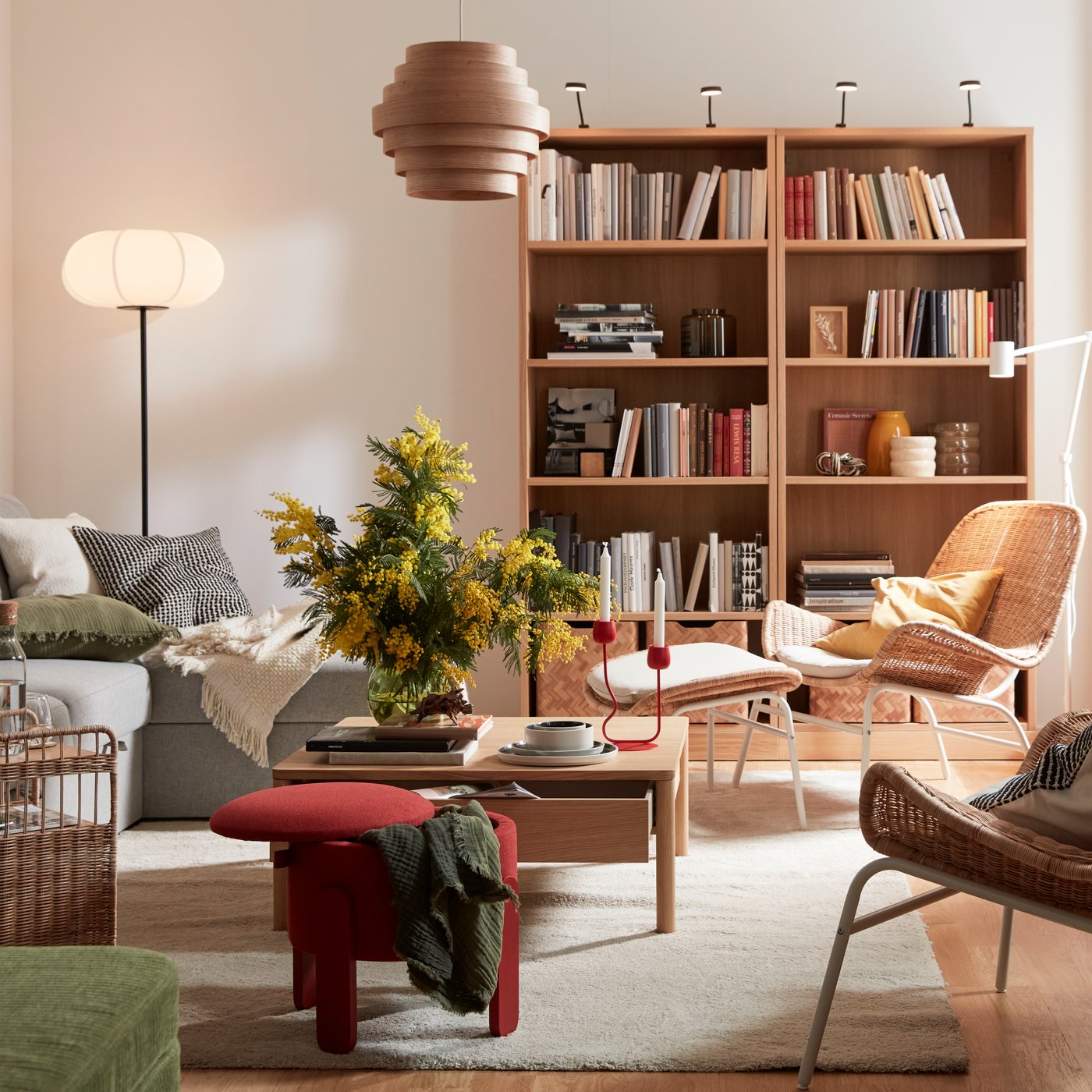 Mysigt vardagsrum med grå soffa, rottingstolar, träbord och bokhyllor. Stora fönster släpper in ljus, gröna växter ger liv åt rummet och lampor skapar mysigt ljus i rummet.