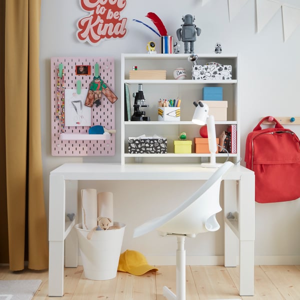 A white PÅHL desk and a white desk chair placed in a children’s room with a shelf and a pink SKÅDIS pegboard.