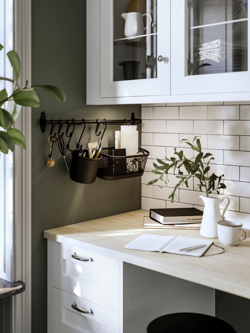 In a kitchen there is a HULTARP rail with hooks and black containers. Next to it, a desk with a jug and a white mug.