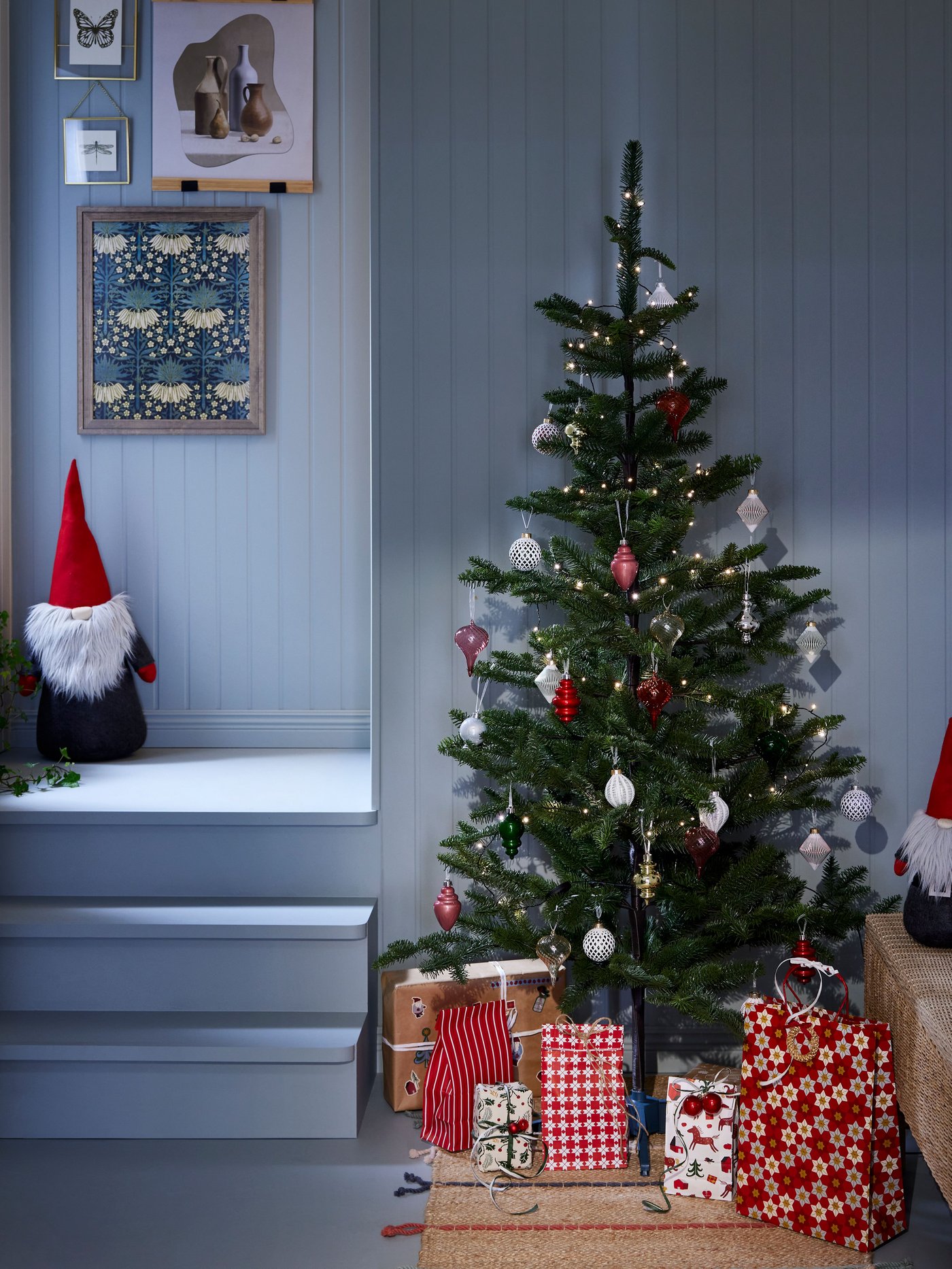 Arbre de nadal decorat amb adorns i boles amb regals sota, i gnom de peluix damunt de l'escala.