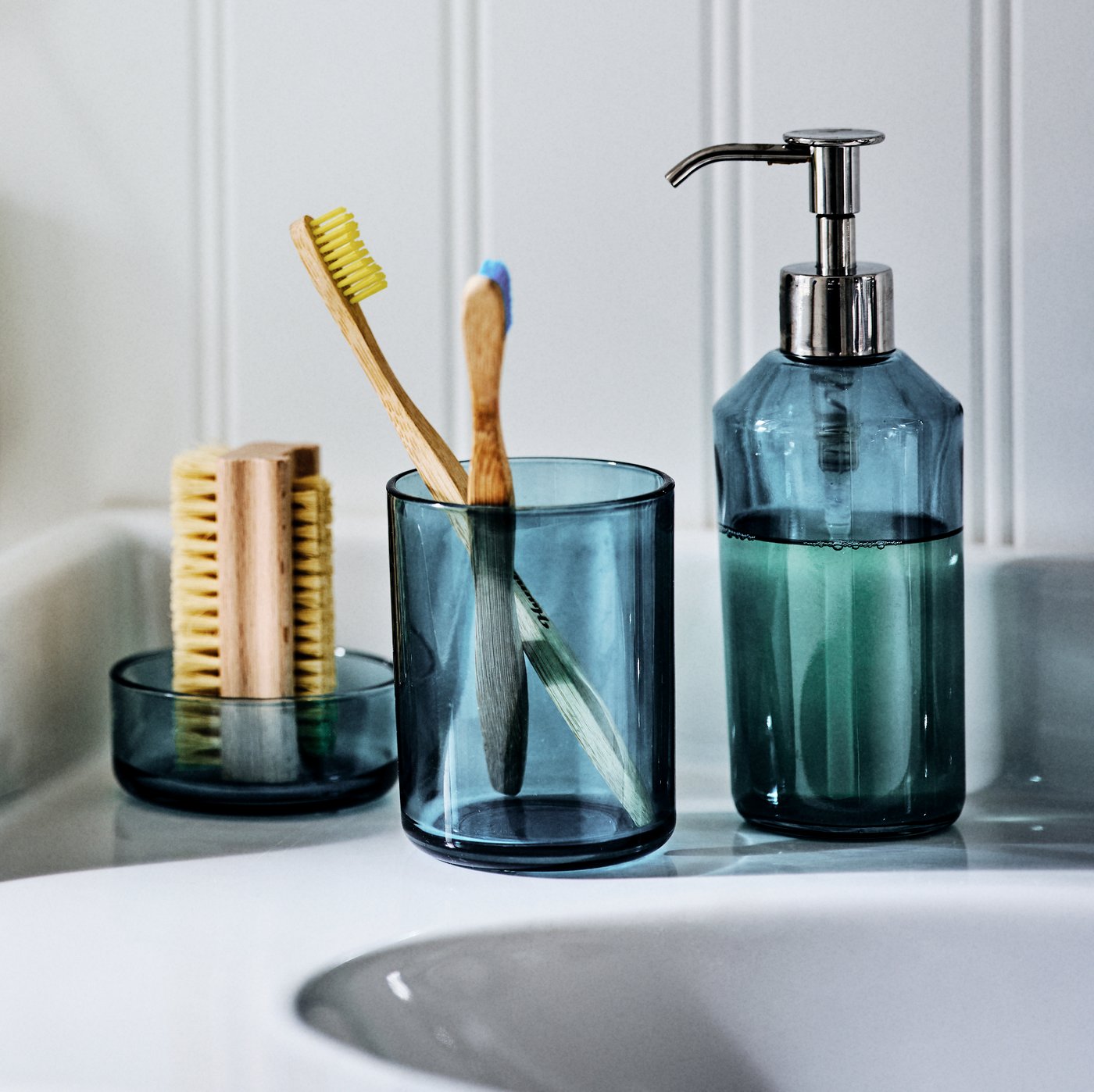 The SKISSEN three-piece glass bathroom set on the side of a sink, holding a nail brush, toothbrushes and liquid soap.