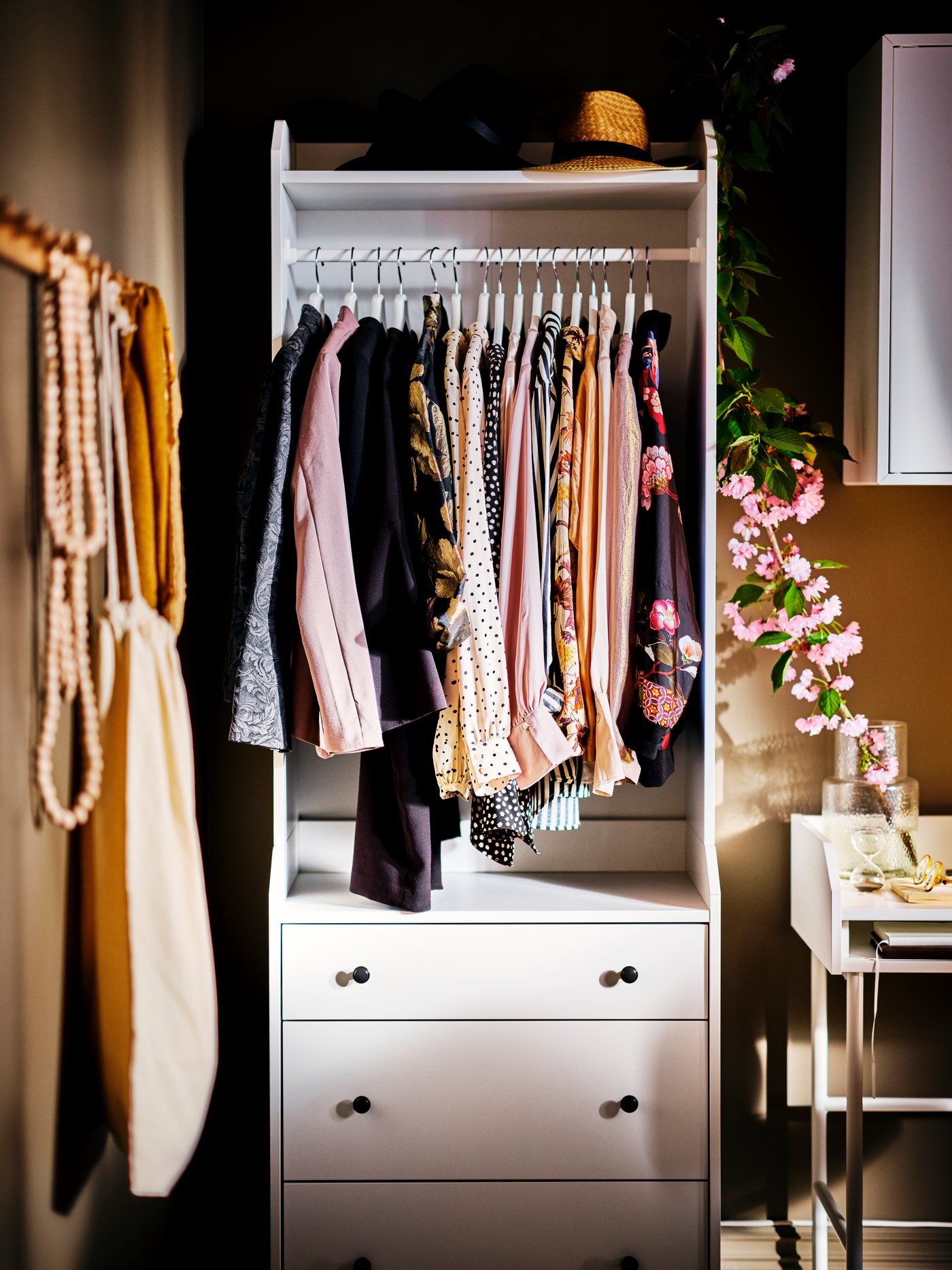 A white HAUGA open wardrobe with 3 drawers with clothes hanging from the rail and flowers in a vase on a desk to the right.