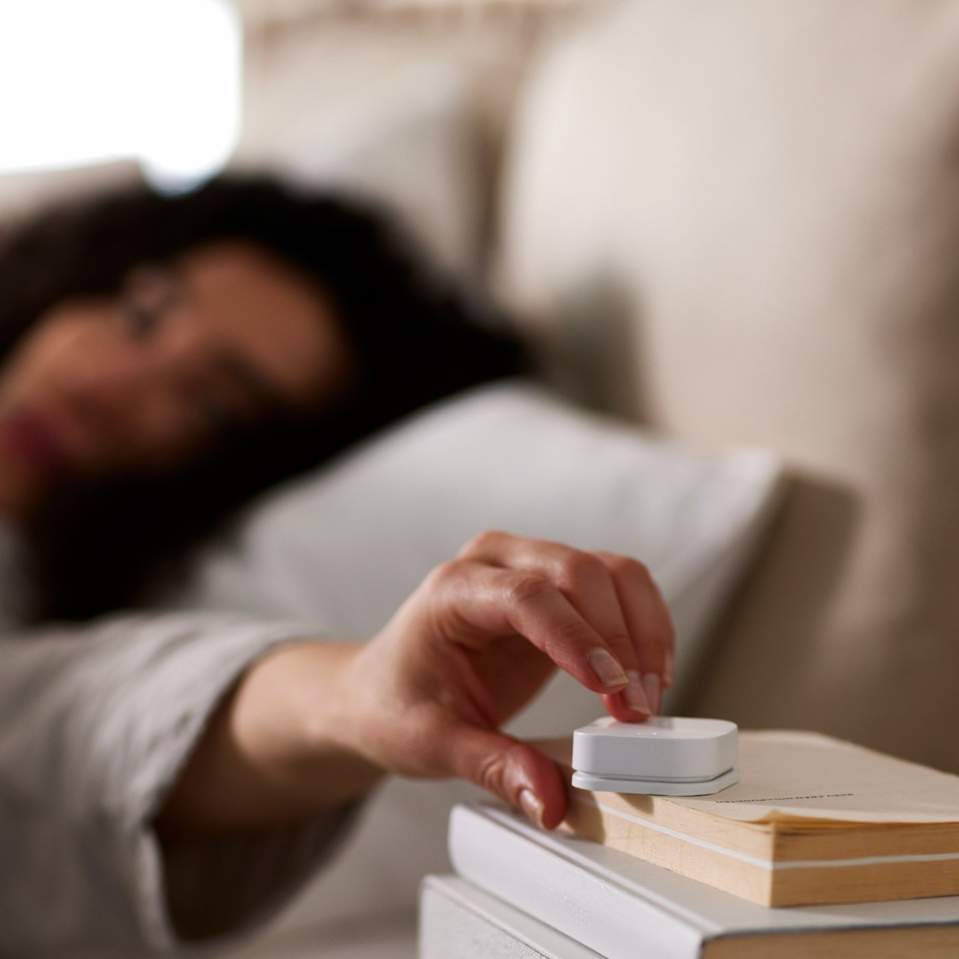 A person lies in bed and reaches out a hand to press a TRÅDFRI wireless dimmer sitting on a pile of books.