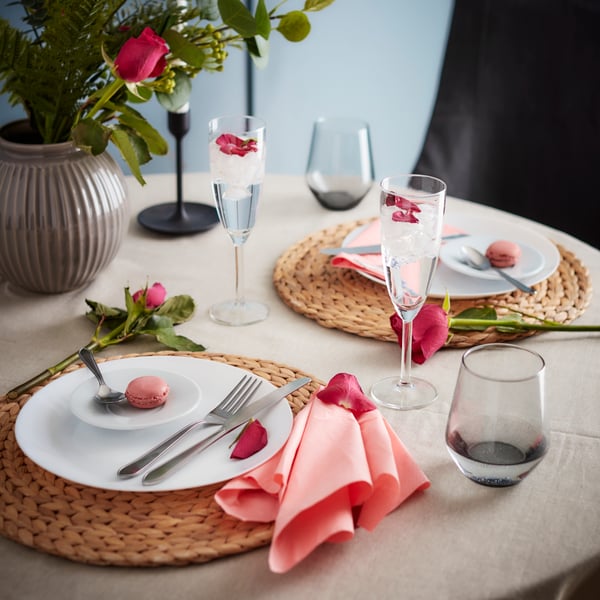 Softly lit dining table with white tablecloth set with white OFTAST dishes for multiple guests. Flowers spill over the side of the table and placed in vases behind.