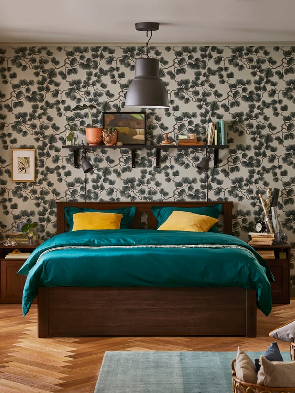 A large grey divan bed with padded headboard, pillows and a quilt in dark blue covers, a beige dress laid on top.