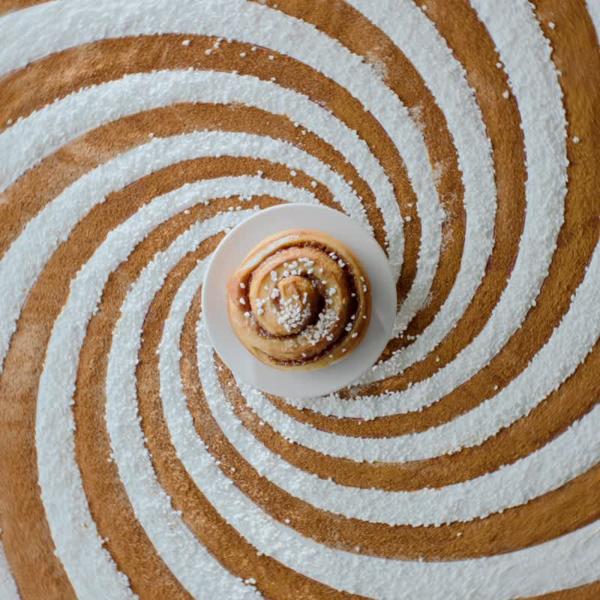 Una brioche alla cannella KAFFEREP vista dall'alto, sopra un motivo a spirale. La brioche e il motivo compiono un movimento rotatorio che crea un effetto ipnotico.
