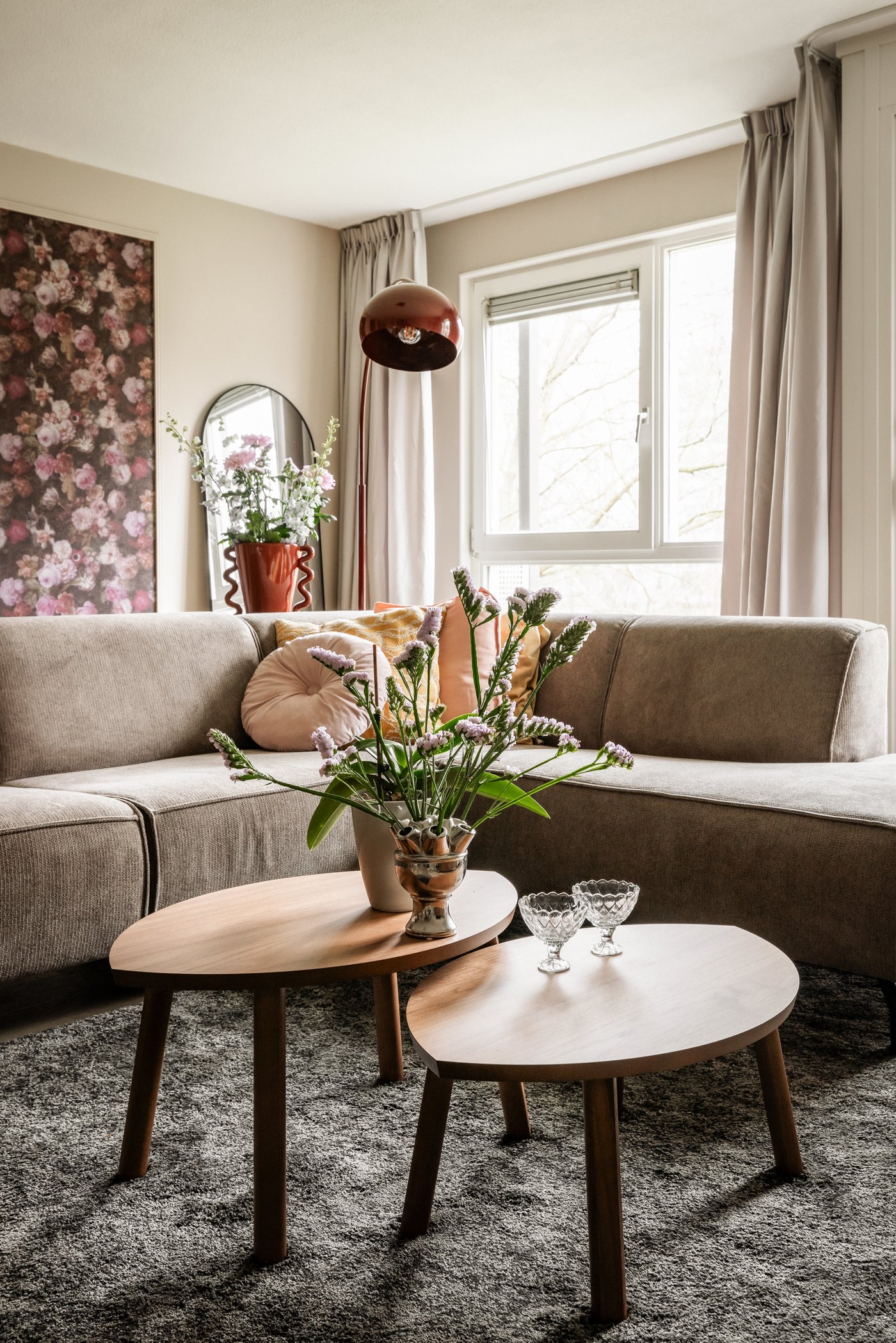Een woonkamer met beige hoekbank die staat op een donkergrijs STOENSE kleed. Achter de bank staat een donkerrode vloerlamp en LINDBYN spiegel. Op de voorgrond staan twee STOCKHOLM bijzettafeltjes in walnootfineer.