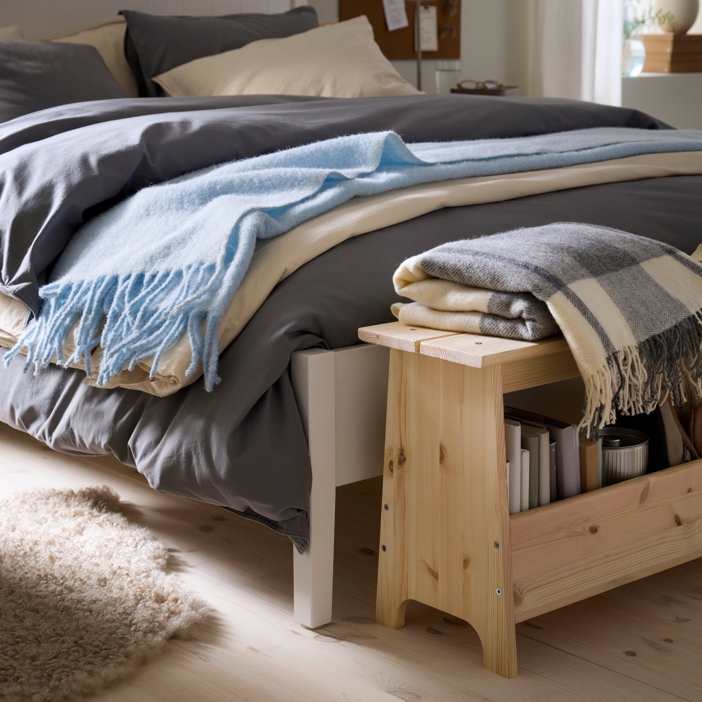 A white bed is layered with duvets and blankets in different shades of blue with a wooden bench at the end.