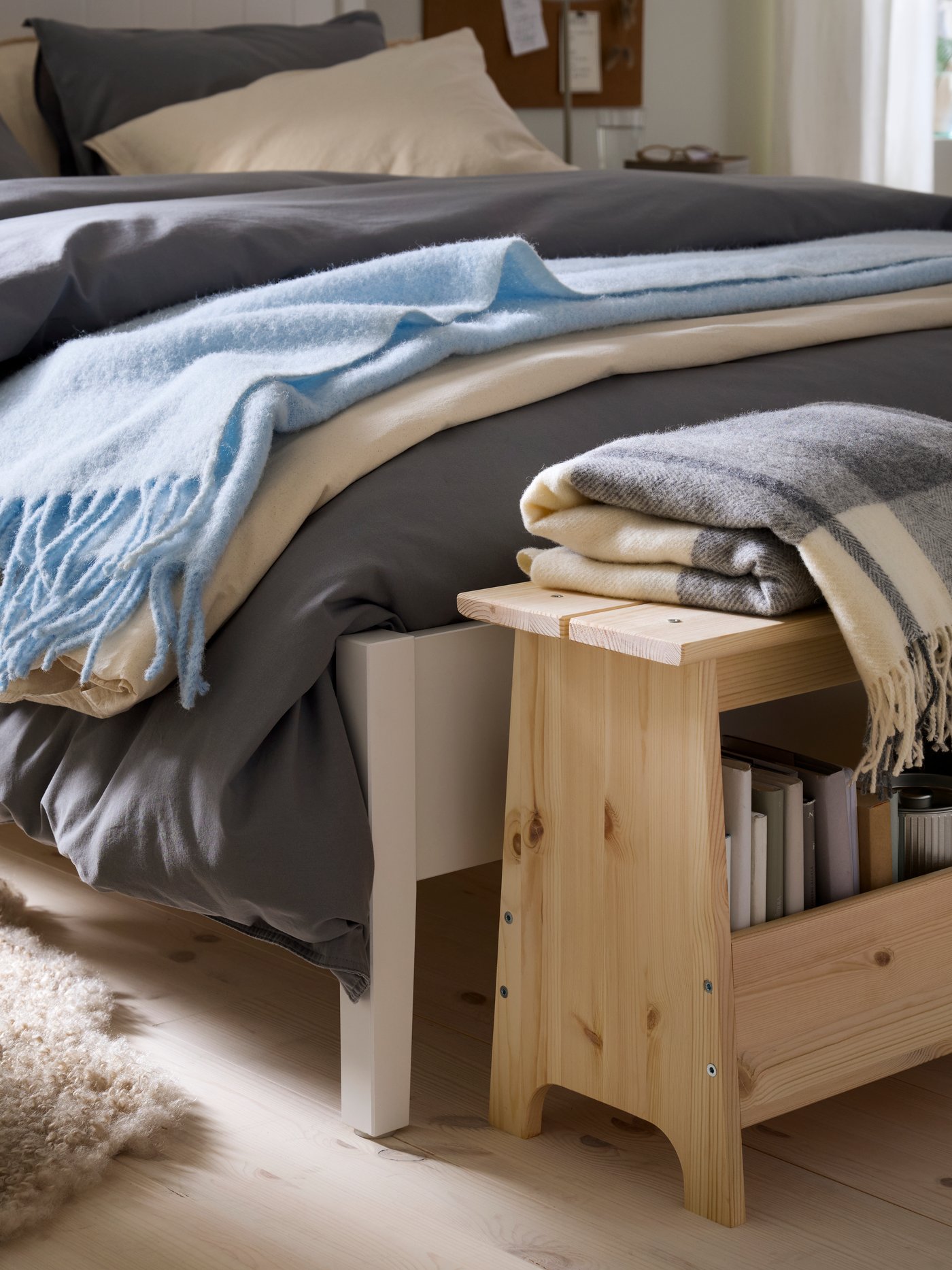 A white bed is layered with duvets and blankets in different shades of blue with a wooden bench at the end.