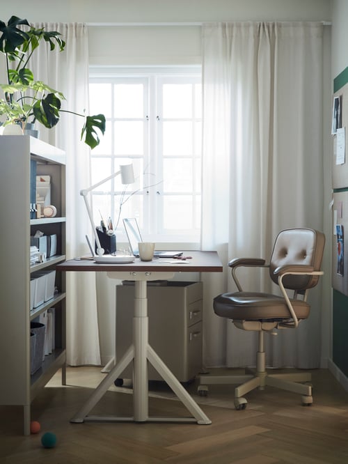 A sit/stand desk by a window with a beige ALEFJÄLL office chair in front of it. A work lamp and laptop is on the desk.