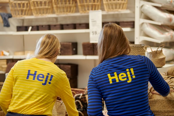 To personer står i er IKEA varhus med ryggen til. De er iført knaldblå og gule trøjer med "Hej!" trykt på bagsiden. 