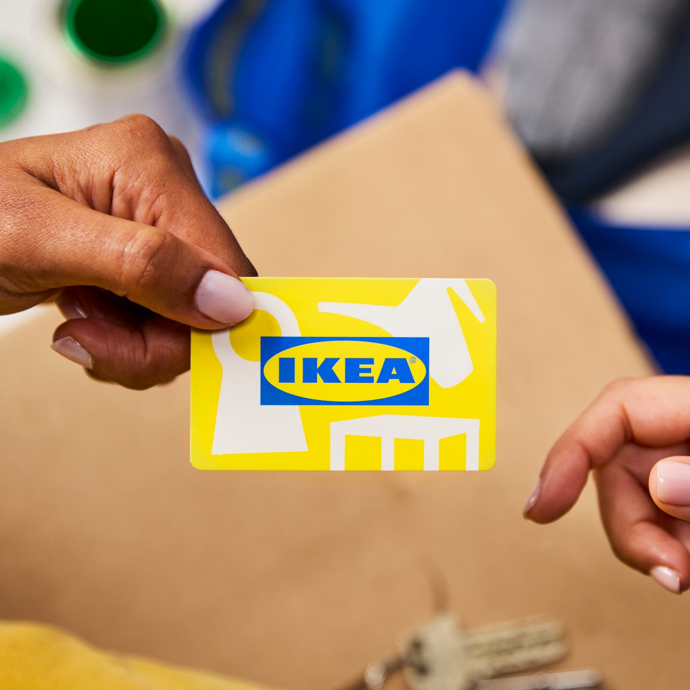 Două mâini schimbă un card cadou IKEA galben cu logo albastru, în fața unui pachet de carton – un gest simplu prin care oferi posibilitatea de a alege tot ce-și dorește pentru casă.