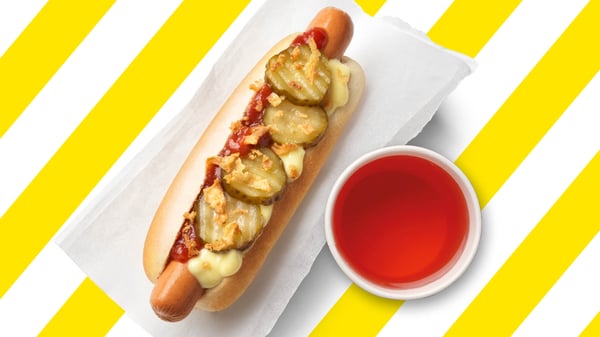 Hot Dog in einem Brötchen mit Röstzwiebeln und Saucen, serviert mit einer Flasche Schwedenkracherl und einem Trinkglas.