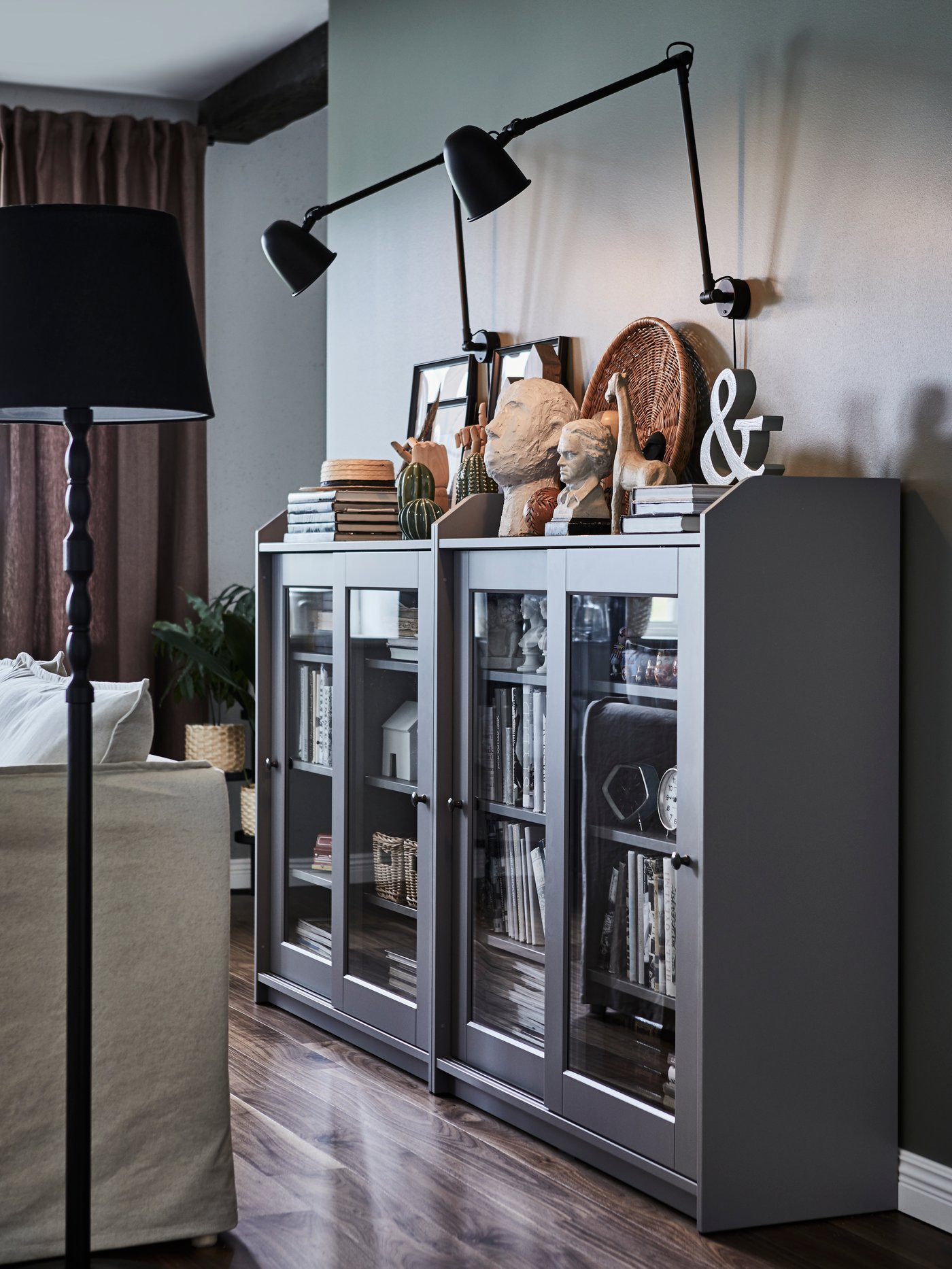 Grey display cabinet with glass doors topped with small statues, books and framed photographs and black anglepoise wall lamps above.