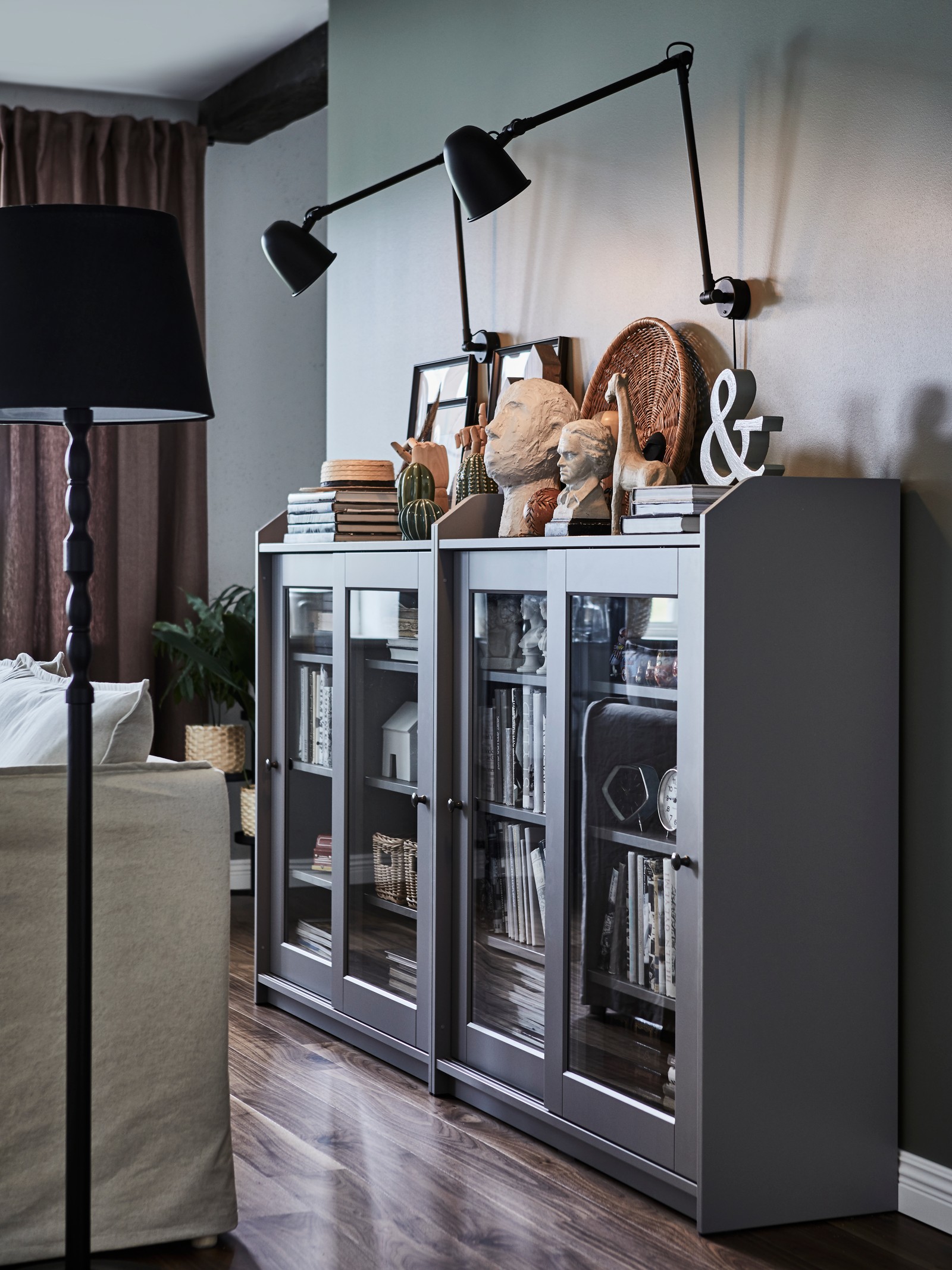 Siva vitrina sa staklenim vratima s malim statuama, knjigama i uramljenim fotografijama i crne zidne lampe iznad.