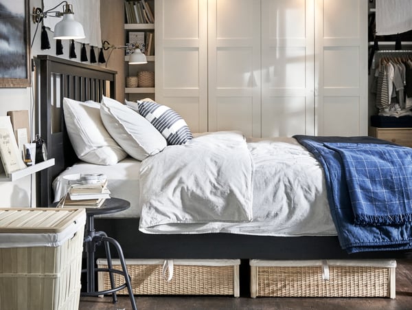 Ein Bett von der Seite, auf dem sich weiße Bettwäsche befindet. Unter dem Bett Aufbewahrungskörbe, hinter dem Bett stehen hohe weiße Schränke.