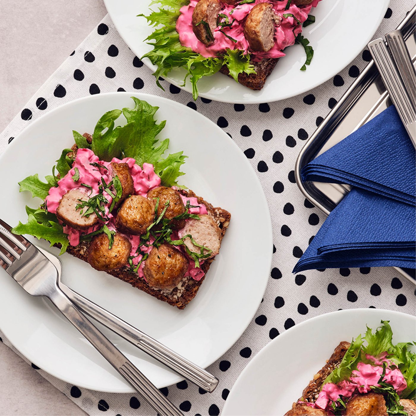 Open roggebrood met gehaktballetjes, roze bietensalade en sla op witte borden, geserveerd op een gestippeld tafelkleed.