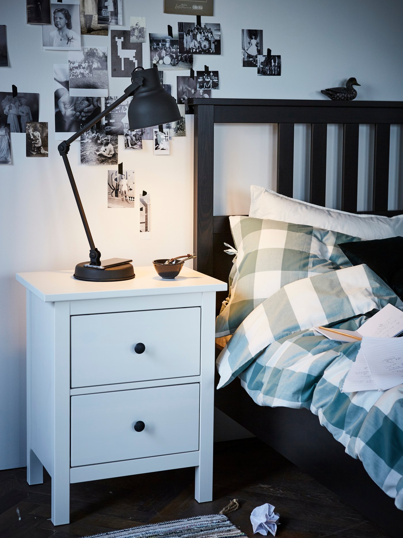 Mesita de noche blanca HEMNES con una lámpara de escritorio negra en su superficie, junto a una cama color negro.