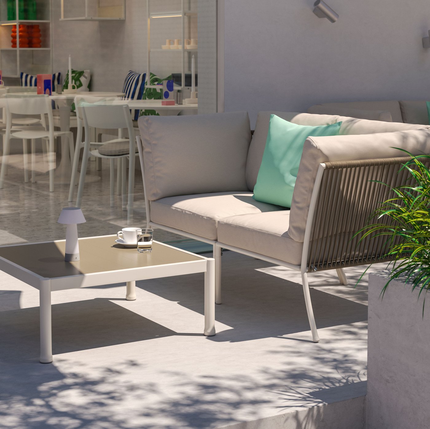 A sunny outdoor café lounging area with a 2-seat white/beige SEGERÖN outdoor conversation set, a blue parasol and greenery.