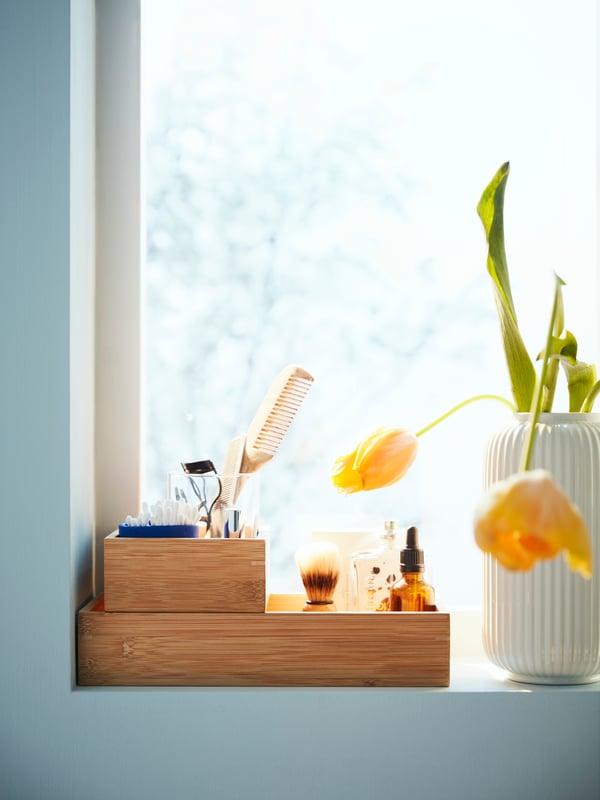 Auf einem Fensterbrett steht ein TAVELAN organizer aus Holz. Darin stehen Deko Sachen und daneben eine Vase mit einer Pflanze darin.