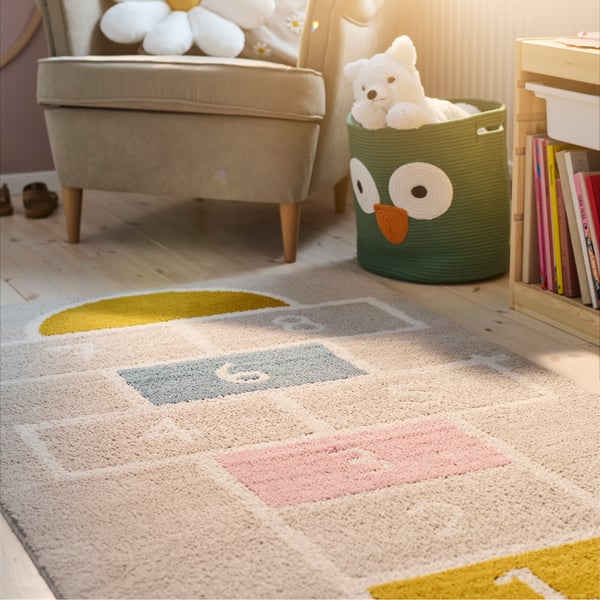 A beige STÄPPSPARV rug on the floor of a sunny children’s bedroom in front of a beige armchair and a basket of toys.
