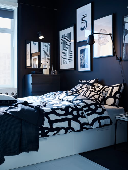 Modern bedroom with black painted wall, bed frame, a quilt cover and a throw next to a chest of six drawers.