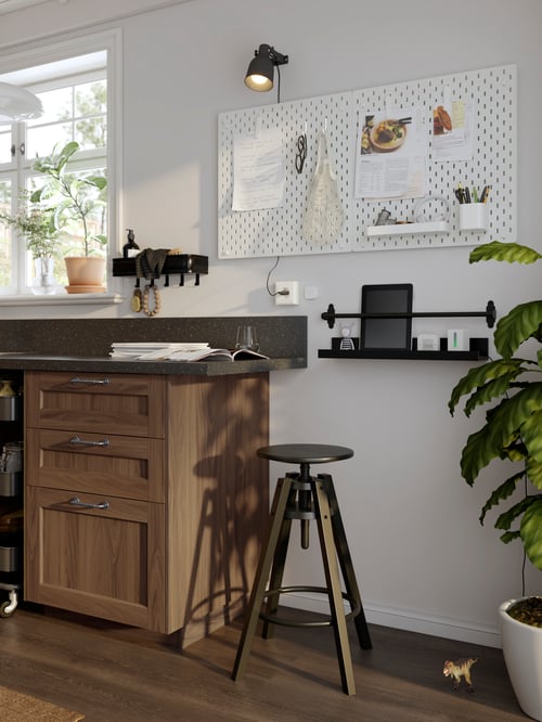 A black DALFRED bar stool at the end of a kitchen counter with a black shelf and white memo board on the wall behind it.
