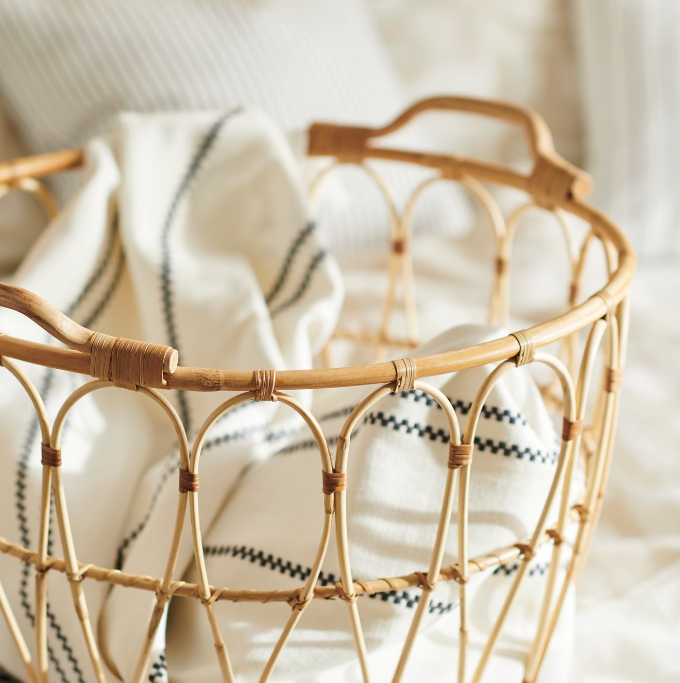 Flätad korg i naturmaterial med handtag, fylld med hopvikta textilier i ljusa färger.