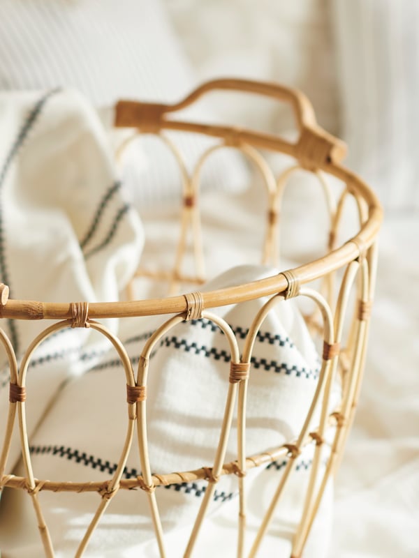 Flätad korg i naturmaterial med handtag, fylld med hopvikta textilier i ljusa färger.