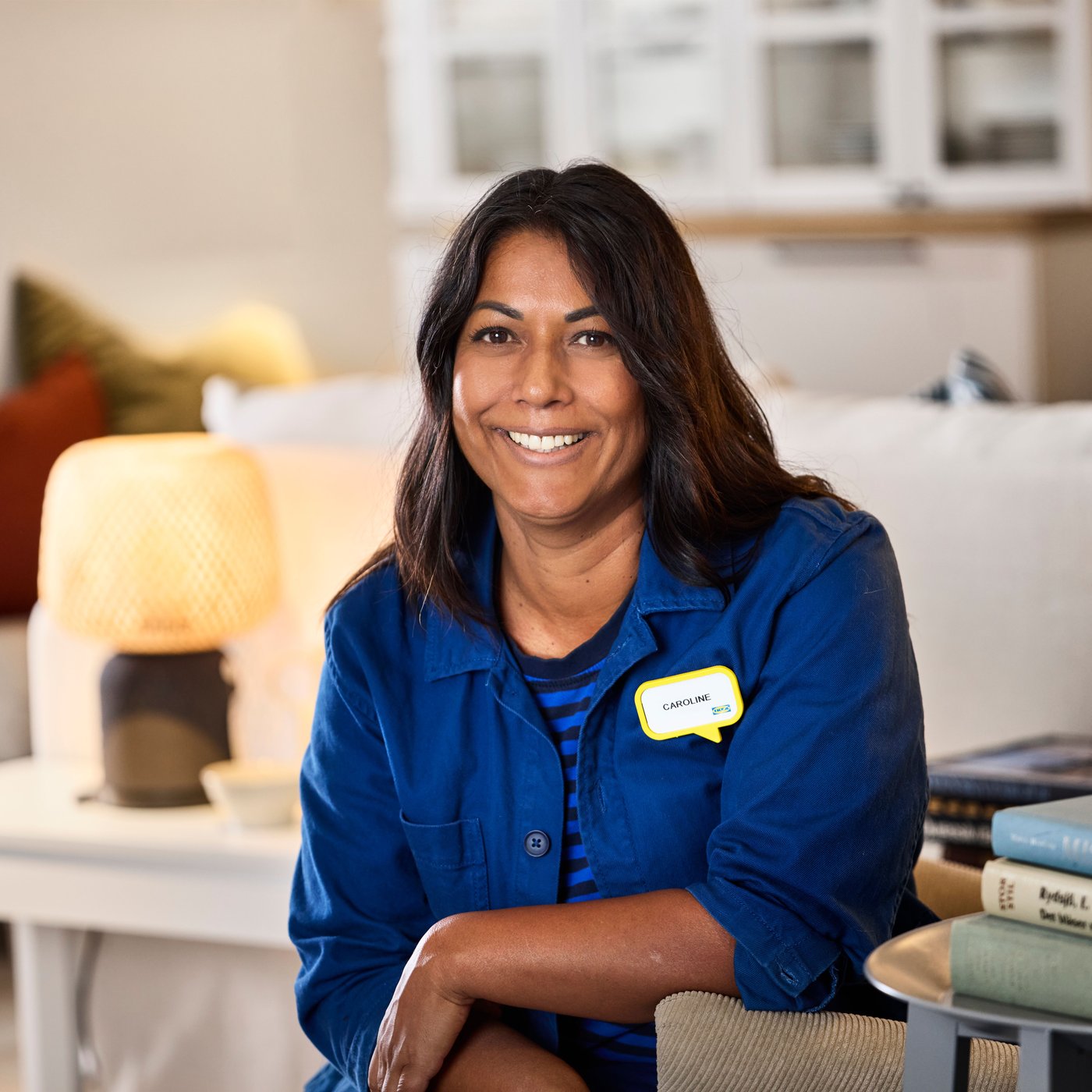 블루 유니폼을 입은 이케아 인테리어 디자이너가 쇼룸에 앉아 카메라를 향해 미소 짓고 있는 모습.