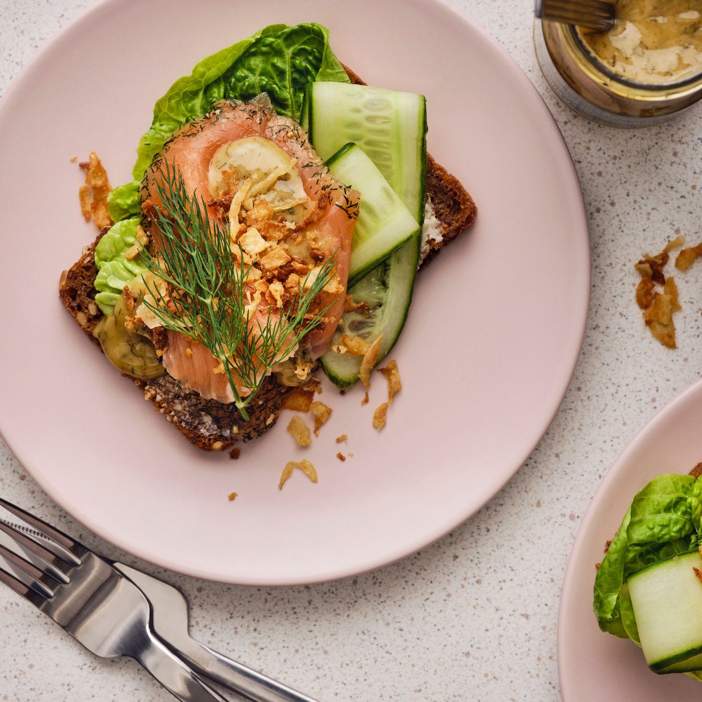 A dark piece of rustic bread with slices of salmon, lettuce, cucumber, dill and a mustard sauce is placed on a pink plate.