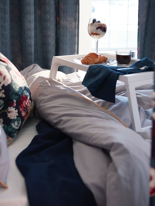 A white, plastic KLIPSK tray with foldable legs holds a luxury breakfast and coffee. It stands on a fluffy bed.