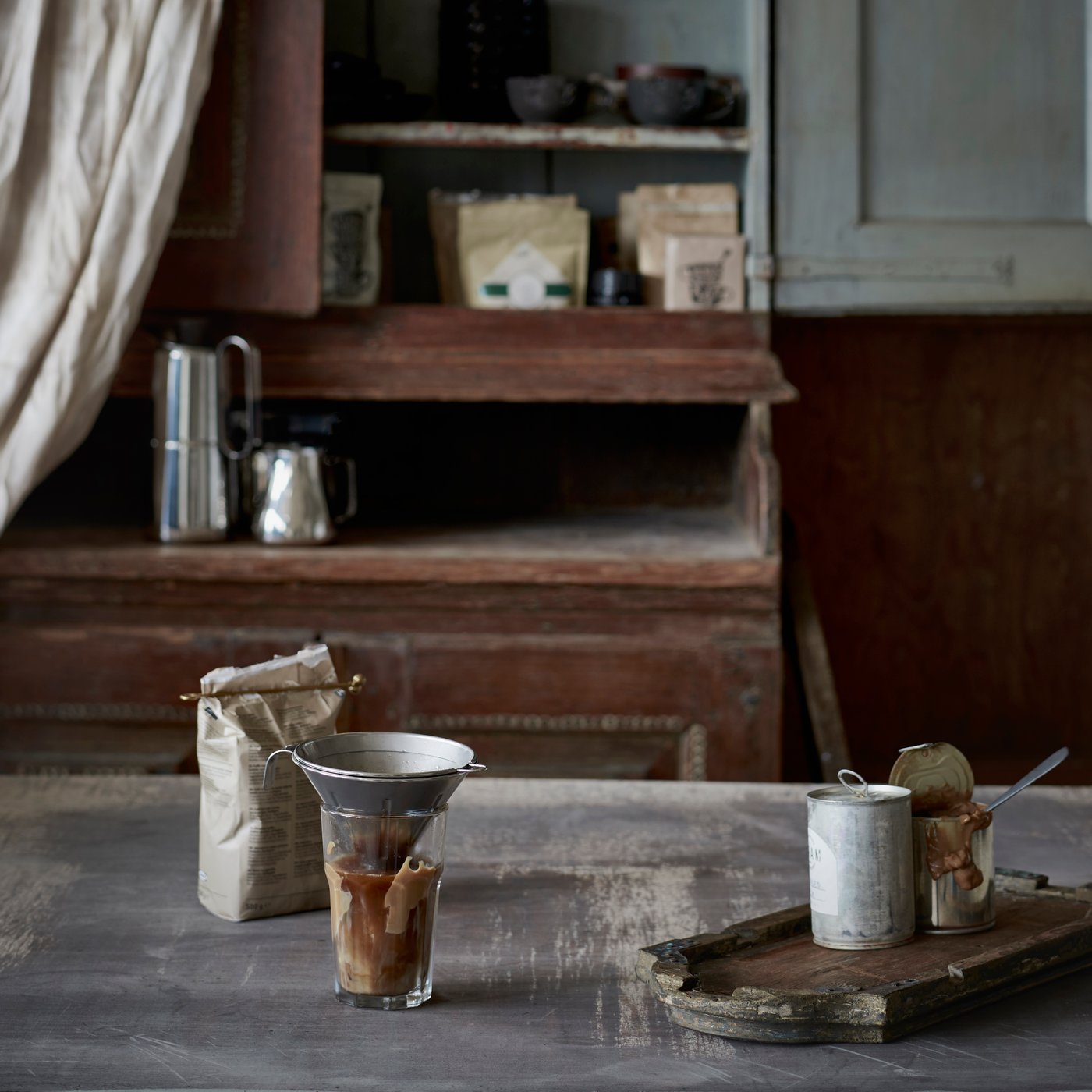 ÖVERST reusable coffee filter in stainless steel on kitchen countertop with iced coffee in glass, coffee beans in linen bag and wooden jug in the background.
