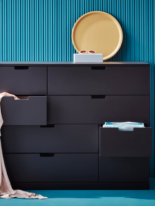 Room with blue wallpaper and blue carpets and dark grey dresser with eight drawers, clothes and decorative tray.