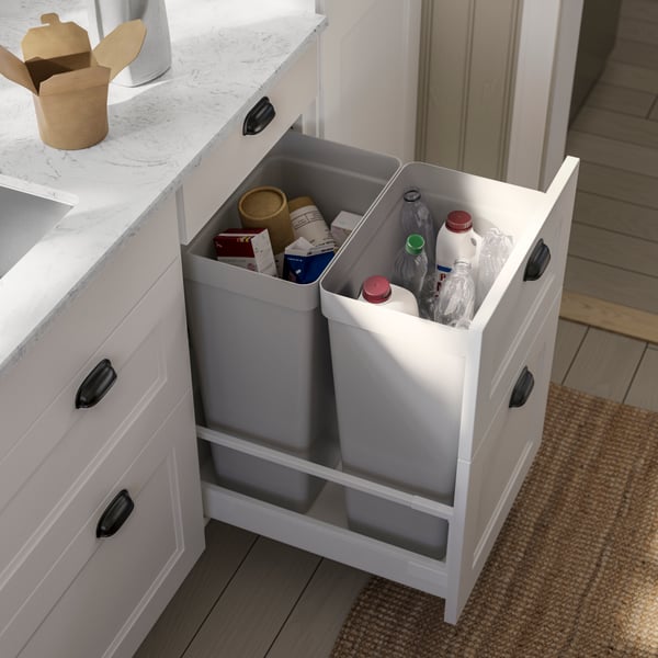 A kitchen featuring a white system with gray countertops, showcasing two gray bins filled with various recyclable containers.