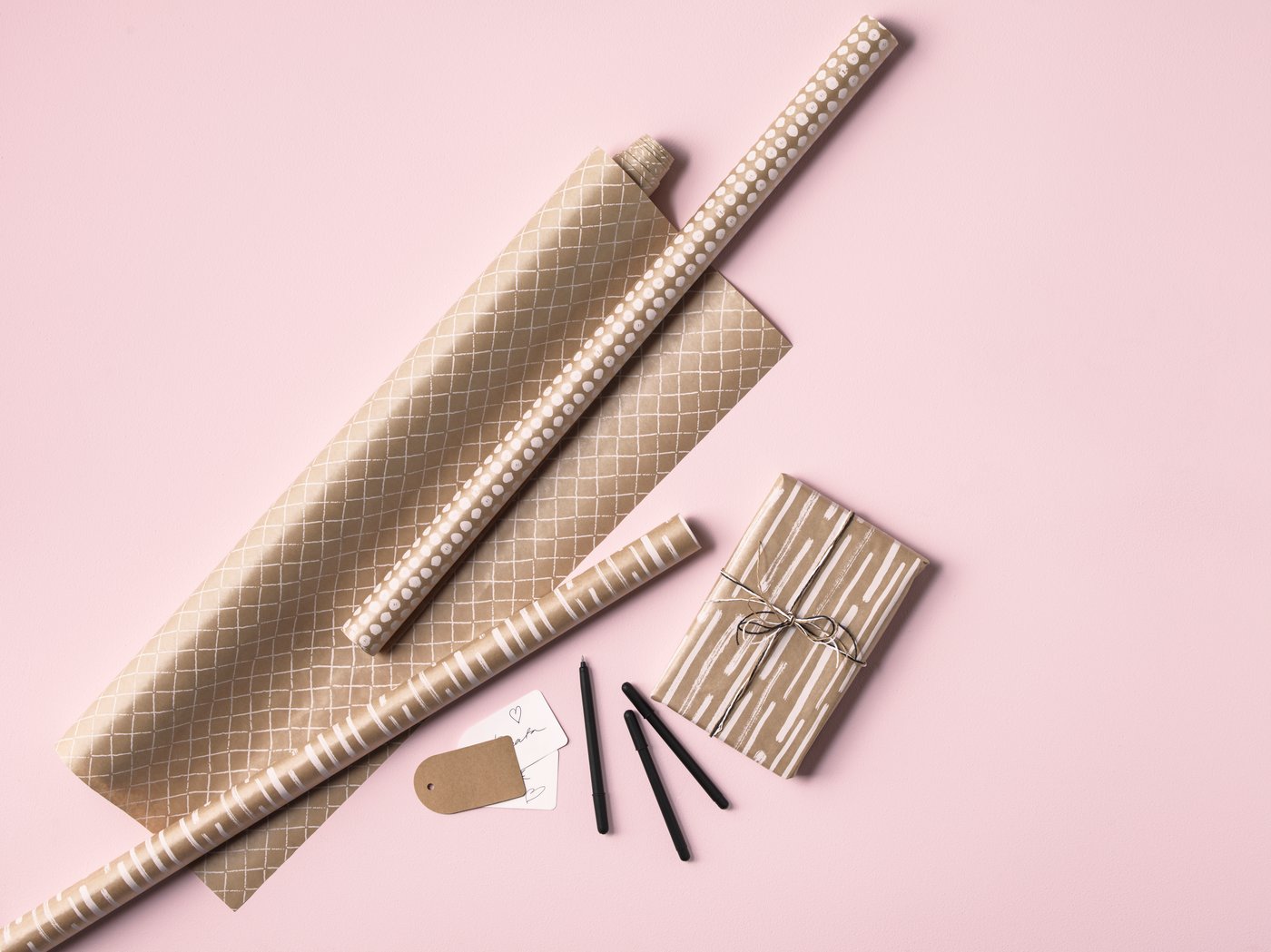 Drei Rollen Geschenkpapier in braun mit unterschiedlichen weißen Mustern auf einem rosa Hintergrund. Daneben Geschenkanhänger, Stifte und ein verpacktes Geschenk. 
