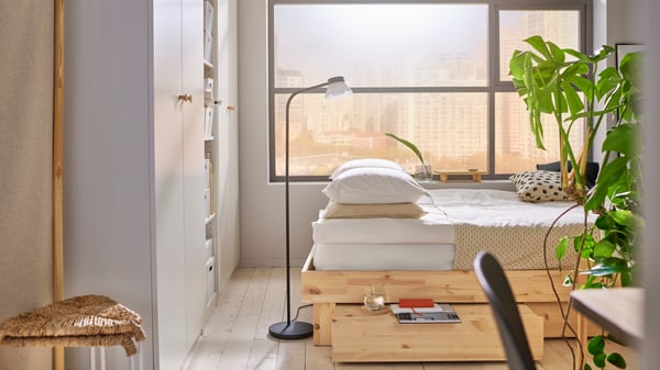 Bedroom with big window, GLAMBERGET bed in untreated wood with drawer underneath, beige bedding, white wardrobes to the left.