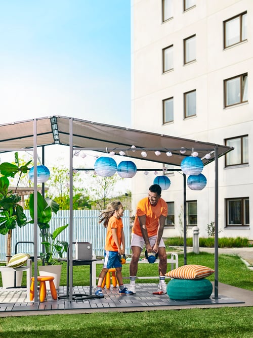 Une arrière-cour estivale, une fille et un père soulevant des poids dans un abri de jardin GUNNÖN avec caillebotis extérieur RUNNEN, des lampes et des plantes.
