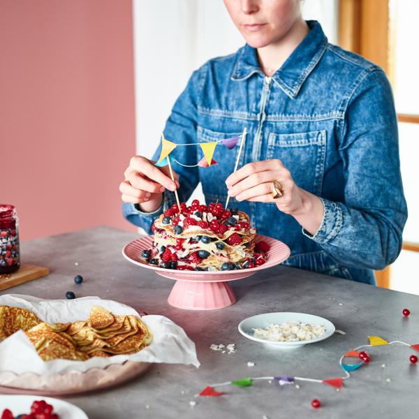 Henkilö tehnyt kerroksellisen kakun PANNKAKOR-letuista, joka on koristeltu erilaisilla marjoilla ja kermavaahdolla.