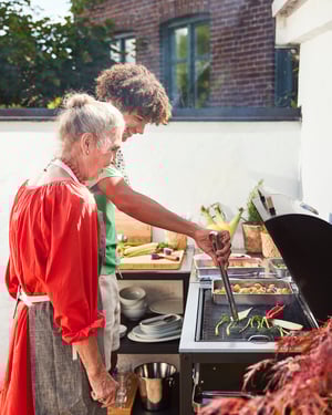 Două persoane gătesc împreună la grătarul într-o bucătărie de exterior, întorcând legume și carne pe plită, într-o curte luminoasă, pregătind o masă relaxată în aer liber.